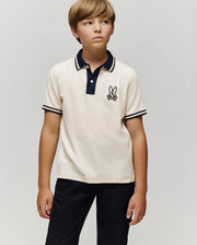 A young boy with light brown hair wears the Psycho Bunny KIDS WADE PIQUE POLO SHIRT (KT0100032) with colorblock tipping and a bunny logo, paired with black pants, standing against a plain light gray background.