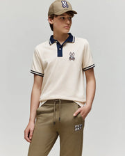 A young person models the Psycho Bunny KIDS WADE PIQUE POLO SHIRT - KT0100032 in beige, featuring colorblock tipping and a cartoon bunny logo, paired with khaki joggers and a matching cap, against a plain light gray background.