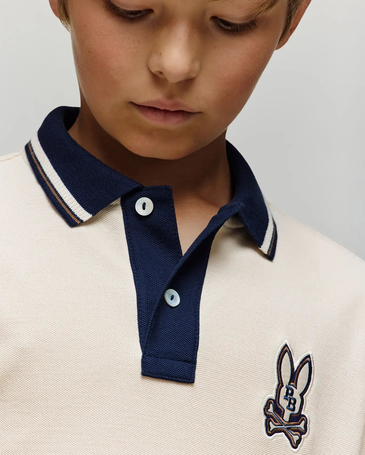 A close-up of a child in the Psycho Bunny Kids Wade Pique Polo Shirt (KT0100032), featuring a navy collar, colorblock tipping, and bunny skull chest patch, looking downward.