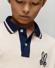A close-up of a child in the Psycho Bunny Kids Wade Pique Polo Shirt (KT0100032), featuring a navy collar, colorblock tipping, and bunny skull chest patch, looking downward.