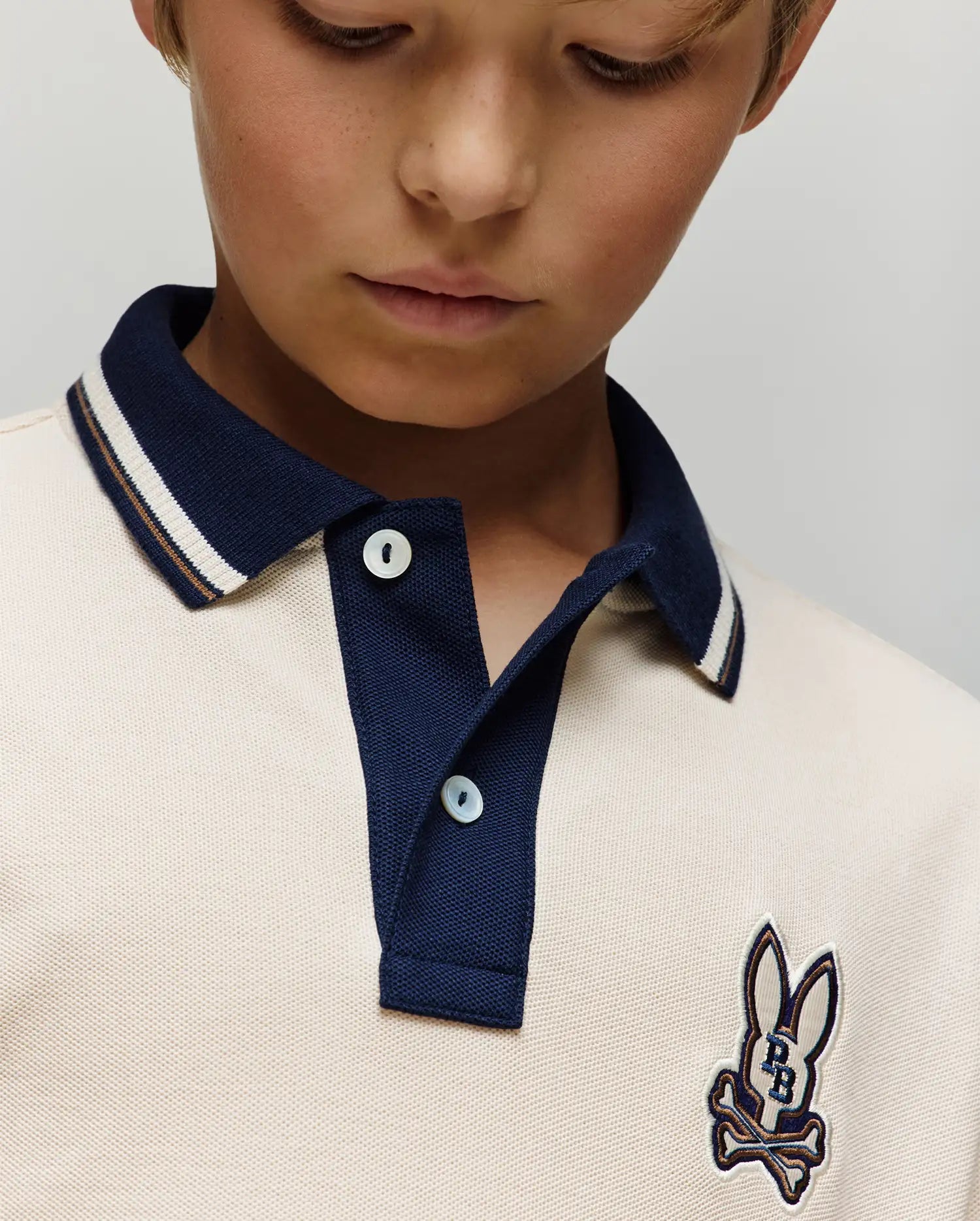 A close-up of a child in the Psycho Bunny Kids Wade Pique Polo Shirt (KT0100032), featuring a navy collar, colorblock tipping, and bunny skull chest patch, looking downward.