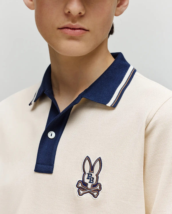 A child wears the Psycho Bunny KIDS WADE PIQUE POLO SHIRT (KT0100032), a cream regular fit Pima cotton polo with navy collar, colorblock tipping, and bunny skull-and-crossbones logo; photo shows lower face and upper torso.