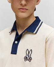 A child wears the Psycho Bunny KIDS WADE PIQUE POLO SHIRT (KT0100032), a cream regular fit Pima cotton polo with navy collar, colorblock tipping, and bunny skull-and-crossbones logo; photo shows lower face and upper torso.