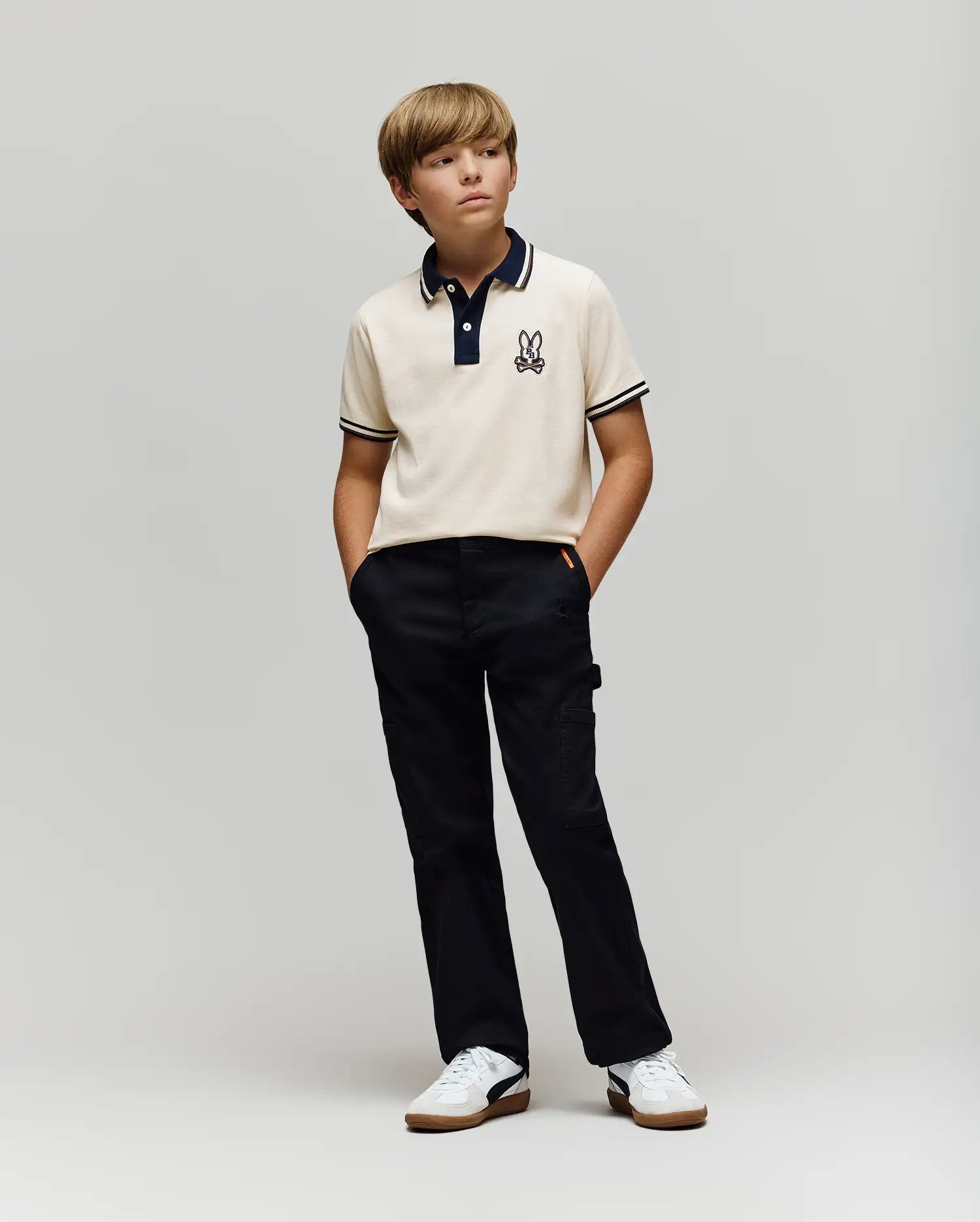 A young boy stands against a plain background, wearing the Psycho Bunny KIDS WADE PIQUE POLO SHIRT (KT0100032) with colorblock tipping and a small bunny logo, paired with black pants and white sneakers. His hands are in his pockets as he looks to the side.