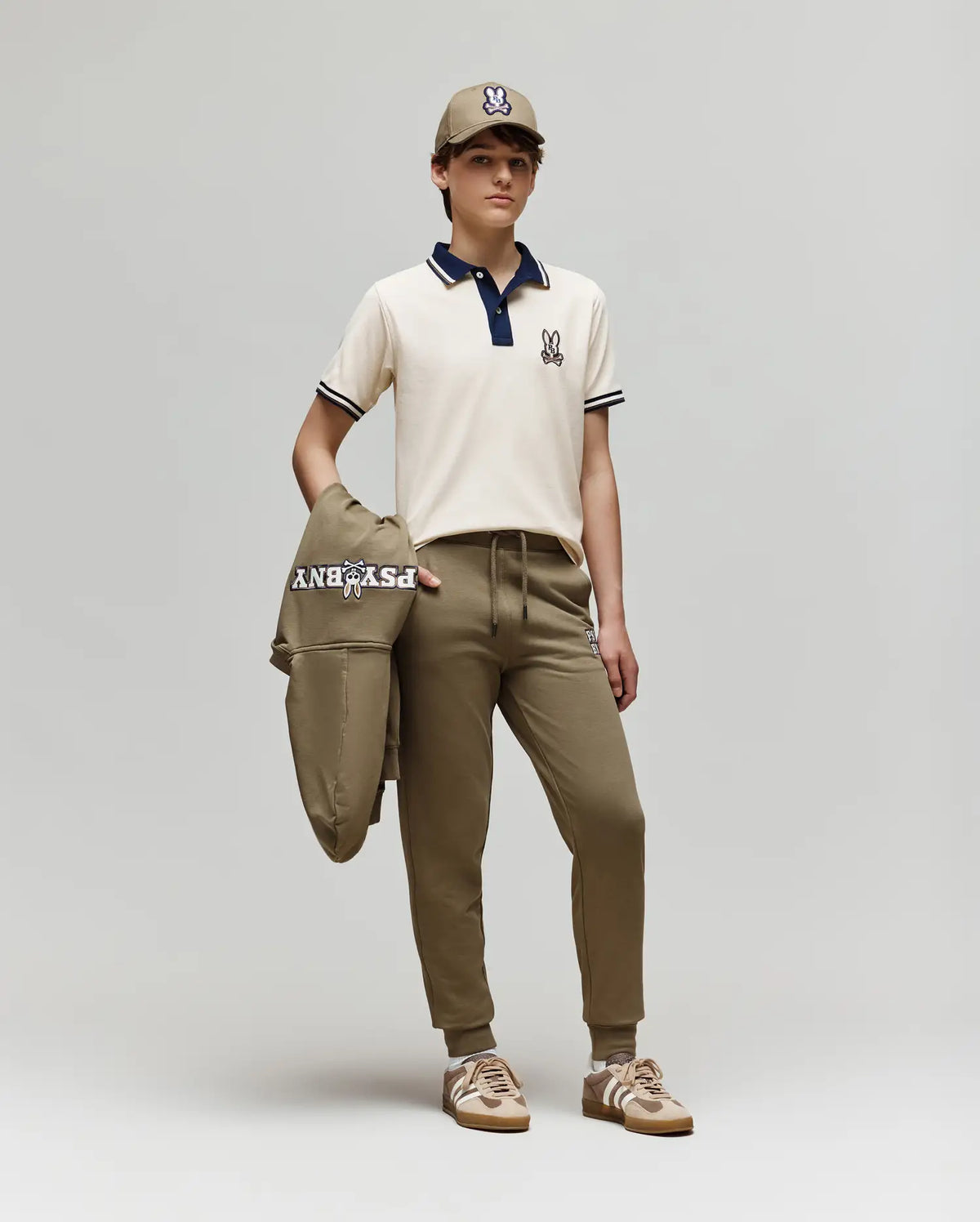 A young person stands against a plain background wearing the Psycho Bunny KIDS WADE PIQUE POLO SHIRT (KT0100032) in off-white, styled with khaki jogger pants, beige sneakers, and holding a matching logoed khaki jacket.