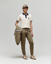 A young person stands against a plain background wearing the Psycho Bunny KIDS WADE PIQUE POLO SHIRT (KT0100032) in off-white, styled with khaki jogger pants, beige sneakers, and holding a matching logoed khaki jacket.