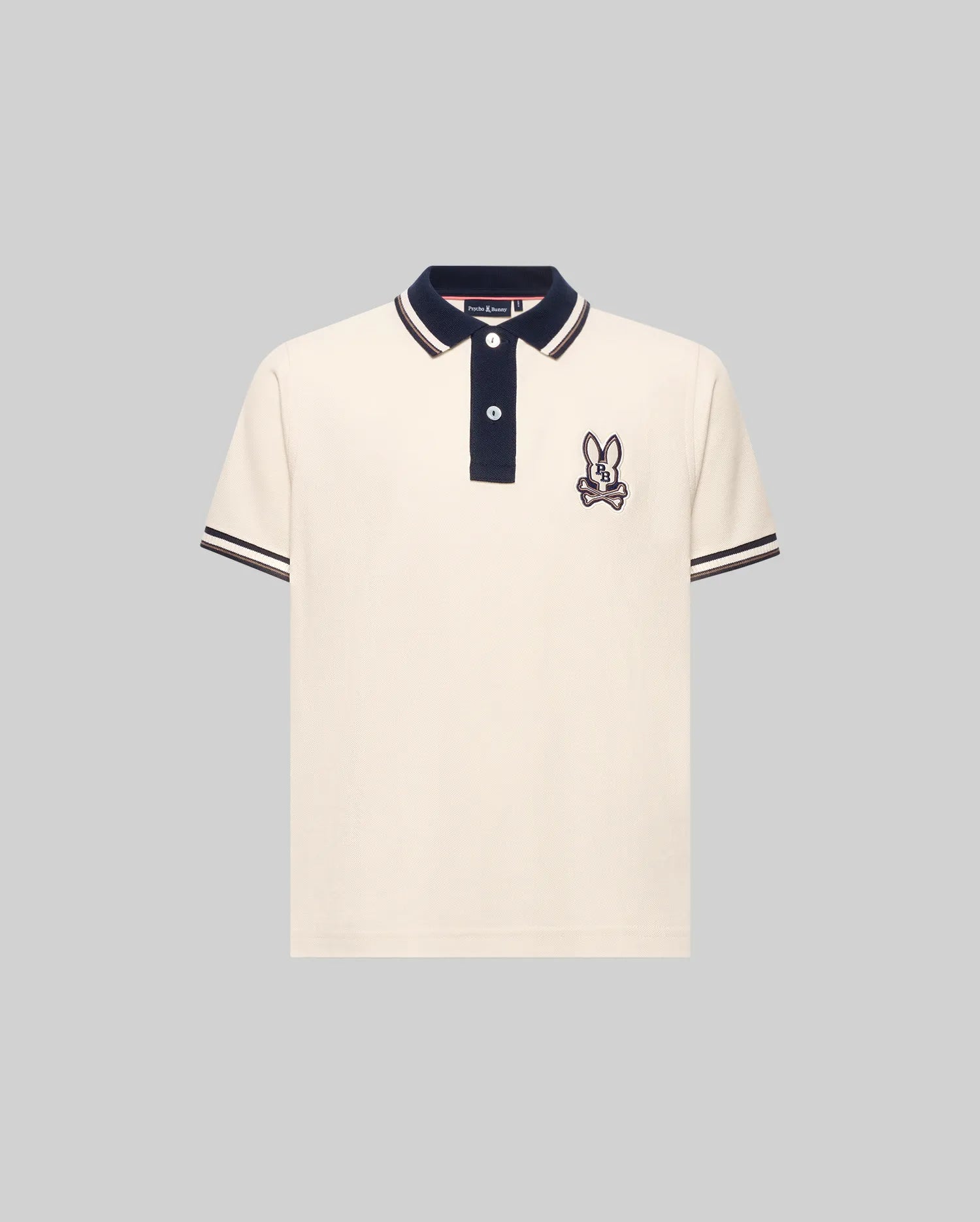 The Psycho Bunny KIDS WADE PIQUE POLO SHIRT (KT0100032) is a regular fit, cream Pima cotton polo with black colorblock tipping, black button placket, and embroidered bunny with crossed bones on the left chest.