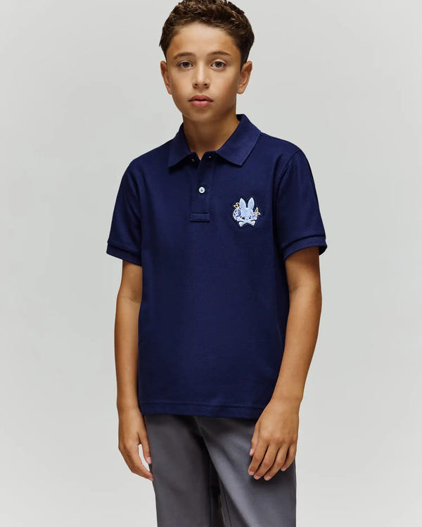 A young boy stands facing forward in a Psycho Bunny KIDS AUGUSTE PIQUE POLO - KT0100033, featuring dark blue Pima cotton with an embroidered crest and chenille logo patch on the chest, paired with gray pants against a plain background.