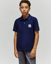 A young boy stands facing forward in a Psycho Bunny KIDS AUGUSTE PIQUE POLO - KT0100033, featuring dark blue Pima cotton with an embroidered crest and chenille logo patch on the chest, paired with gray pants against a plain background.