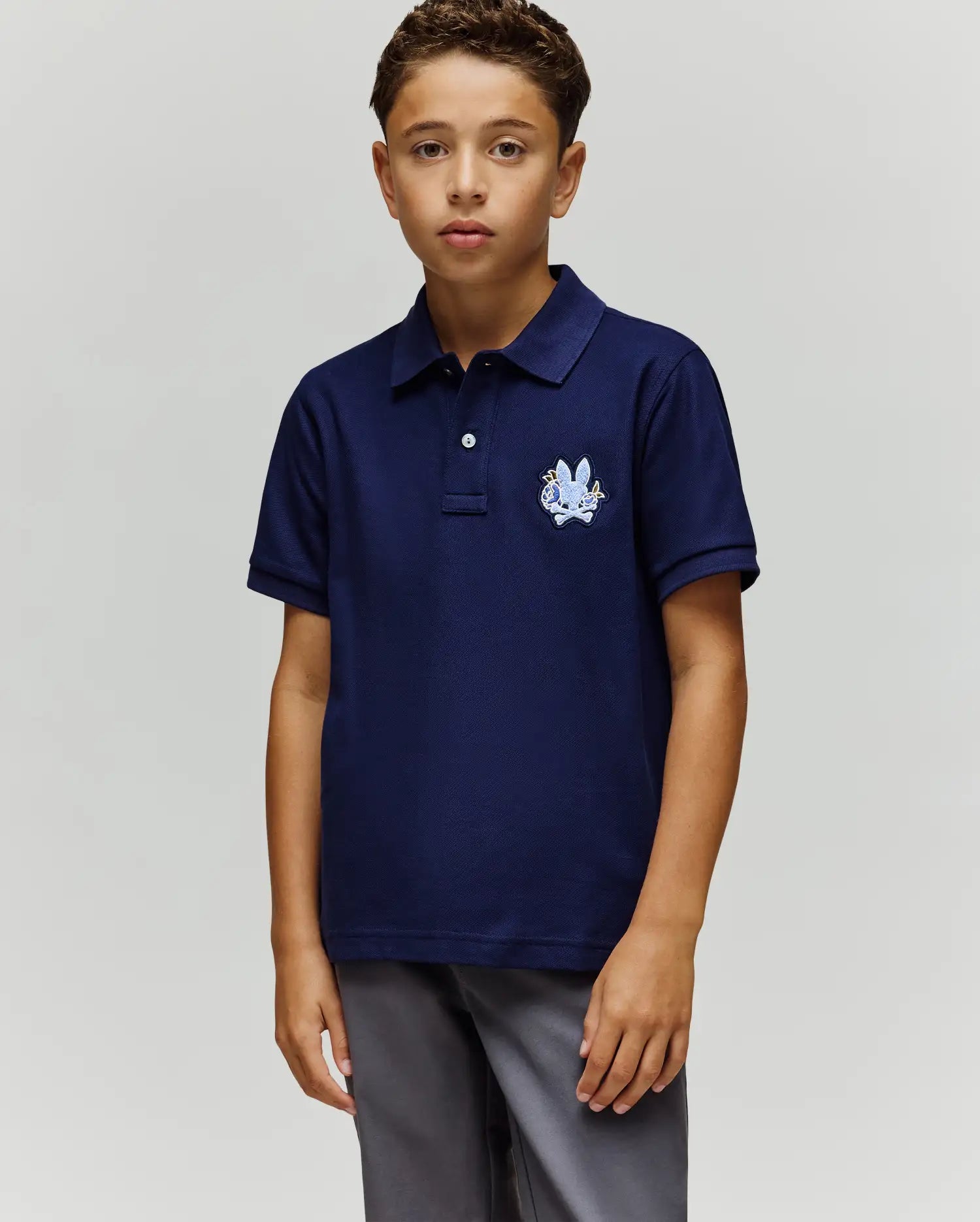 A young boy stands facing forward in a Psycho Bunny KIDS AUGUSTE PIQUE POLO - KT0100033, featuring dark blue Pima cotton with an embroidered crest and chenille logo patch on the chest, paired with gray pants against a plain background.
