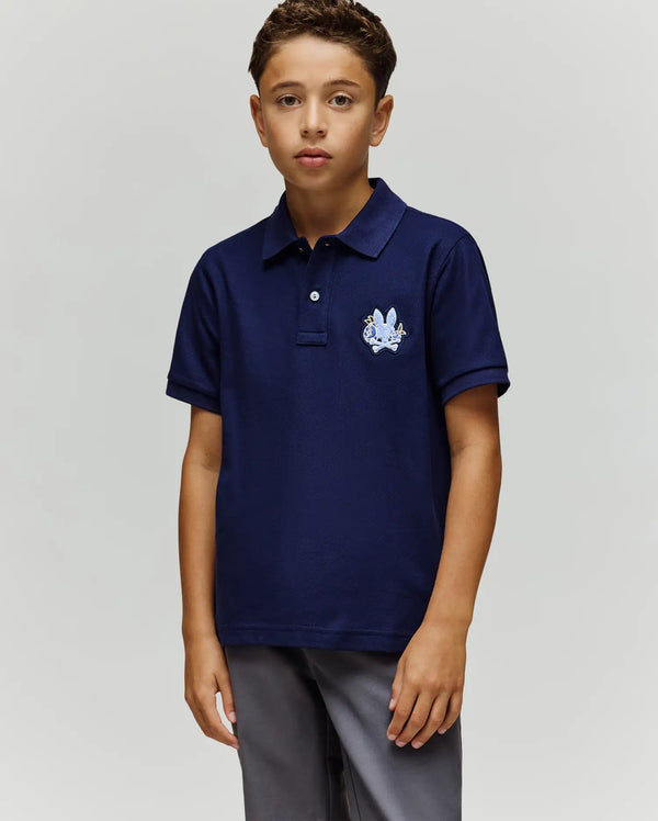 A young boy stands facing forward in a Psycho Bunny KIDS AUGUSTE PIQUE POLO - KT0100033, featuring dark blue Pima cotton with an embroidered crest and chenille logo patch on the chest, paired with gray pants against a plain background.