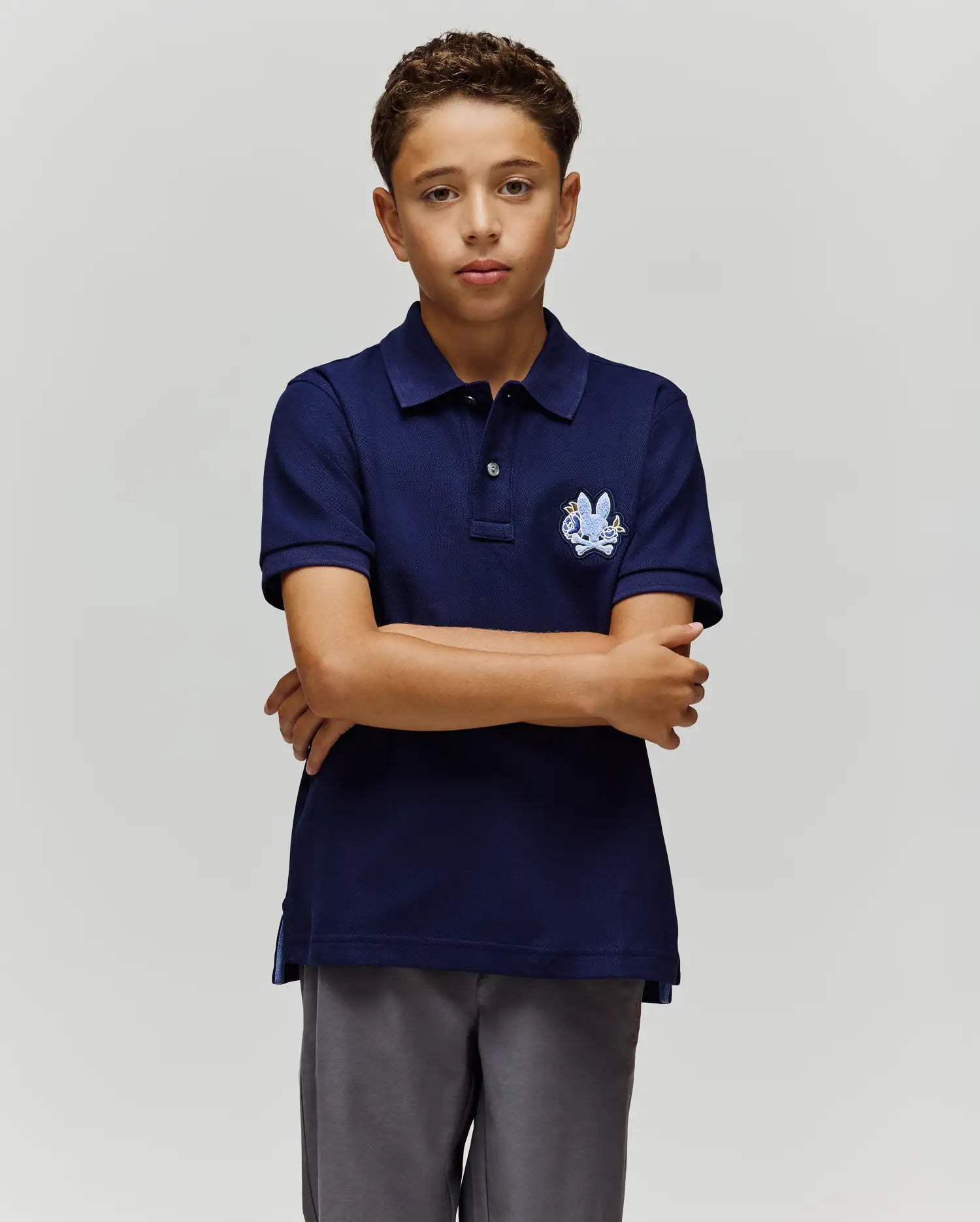 A boy with short curly hair stands with arms crossed, wearing the Psycho Bunny KIDS AUGUSTE PIQUE POLO (KT0100033) in navy blue, featuring a chest crest and gray pants, set against a plain light gray background.