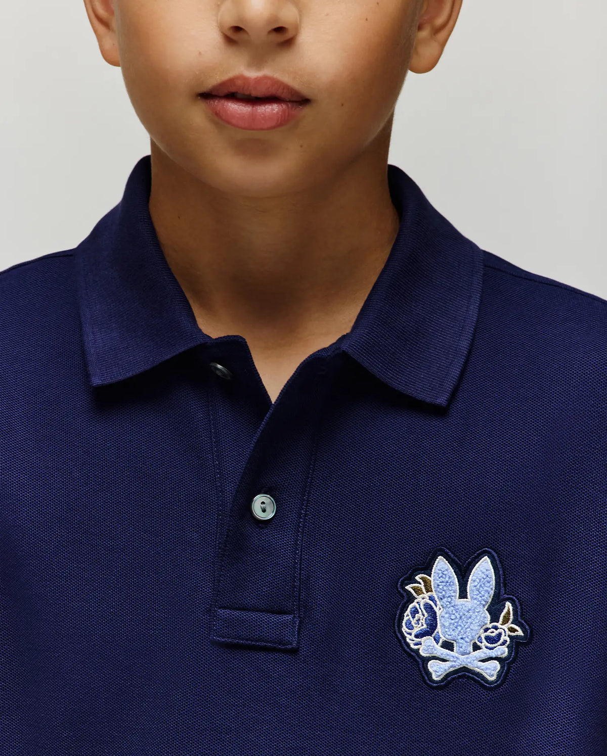 A close-up of a child in the Psycho Bunny KIDS AUGUSTE PIQUE POLO (KT0100033), a navy blue Pima cotton polo featuring a light blue cartoon bunny and crossbones chenille logo patch on the left chest. Only the lower half of the child’s face is visible.
