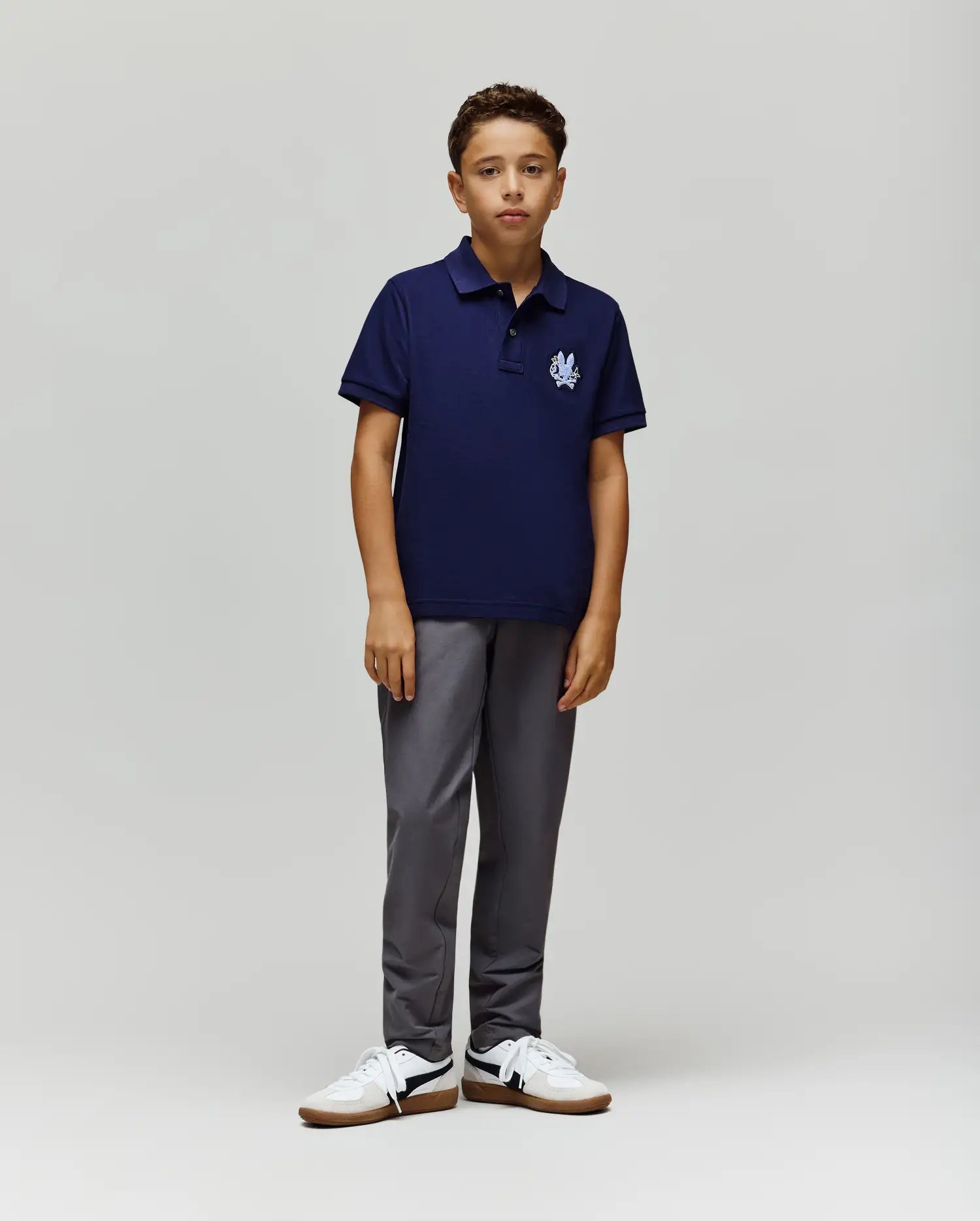 A boy stands against a light background, wearing the Psycho Bunny KIDS AUGUSTE PIQUE POLO (KT0100033) in navy blue with a small white emblem, paired with gray pants and white sneakers. He faces forward with a neutral expression.