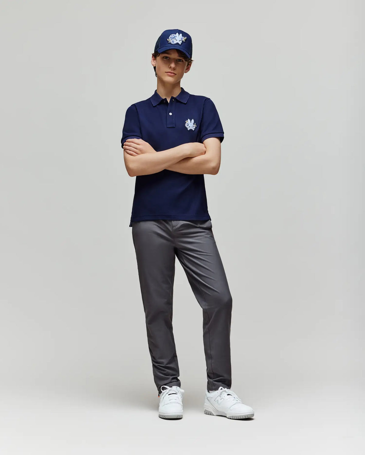 A young person stands with arms crossed against a plain light background, wearing a navy polo, gray pants, white sneakers, and the Psycho Bunny KIDS AUGUSTE TRUCKER CAP (KO1500022) with a bold white chenille patch logo.