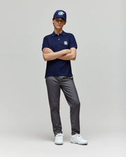 A young person stands with arms crossed against a plain light background, wearing a navy polo, gray pants, white sneakers, and the Psycho Bunny KIDS AUGUSTE TRUCKER CAP (KO1500022) with a bold white chenille patch logo.