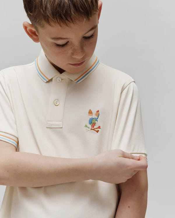 Kids Ara Pique Polo in cream, featuring a classic fit and pique texture. It has a small, multi-colored embroidered Psycho Bunny skull logo on the left chest, with blue and orange striped collar and cuffs.