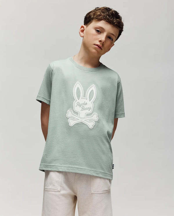 A boy with short brown hair wears the Psycho Bunny KIDS DEREK CENTER GRAPHIC TEE (KT0200006)—a regular fit, light green Pima cotton T-shirt with an embroidered bunny skull and crossbones—paired with light shorts, standing against a gray background.