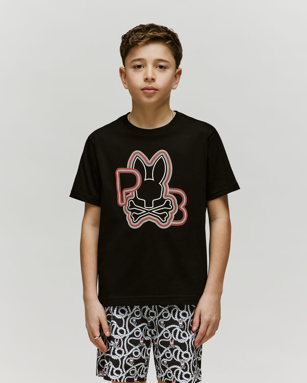 A boy faces the camera in a Psycho Bunny KIDS CARLO GRAPHIC TEE (KT0200009), a black crewneck made from soft Pima cotton, featuring a large bunny skull logo, paired with patterned black-and-white shorts, against a plain background.