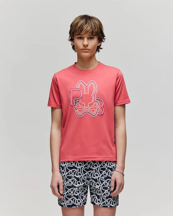 Teenage boy with straight brown hair wears the Psycho Bunny KIDS CARLO GRAPHIC TEE (KT0200009) featuring a stylized bunny and bones logo, paired with black-and-white patterned shorts, standing against a plain light gray background.