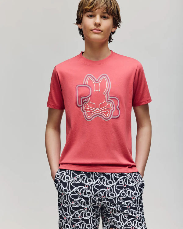 Teen boy with brown hair wears the Psycho Bunny KIDS CARLO GRAPHIC TEE (KT0200009), a red shirt with a stylized bunny and skull design, paired with black-and-white patterned shorts, hands in pockets against a plain light background.