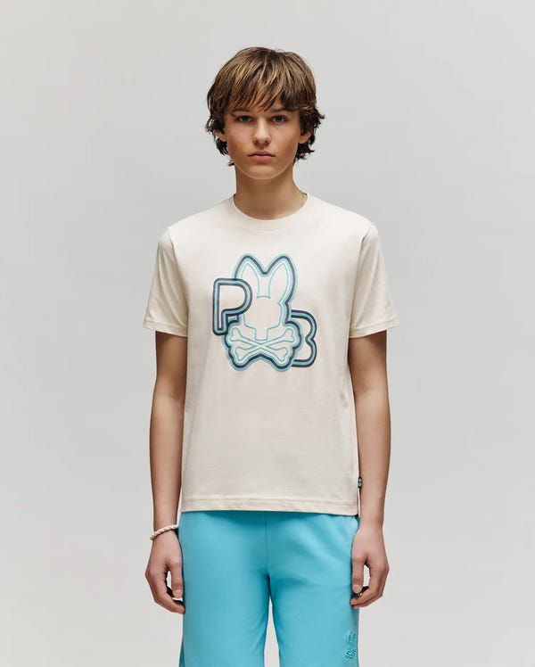 A young person faces forward against a light gray background, wearing Psycho Bunny’s KIDS CARLO GRAPHIC TEE (KT0200009) in cream with a blue bunny and crossbones graphic, paired with light blue shorts.