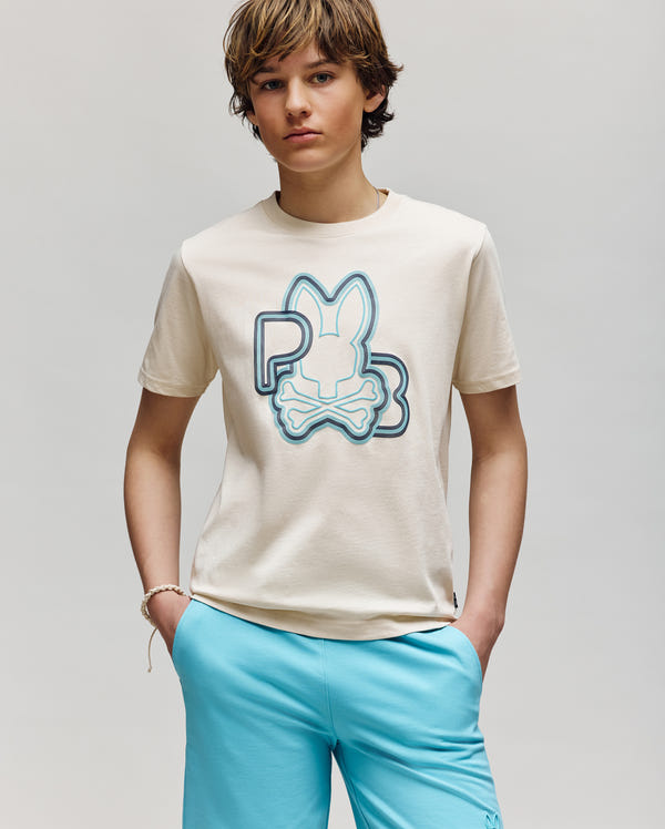A young person with brown hair stands against a plain background, wearing the Psycho Bunny KIDS CARLO GRAPHIC TEE (KT0200009) in beige with a large blue bunny and "PB" design, paired with light blue shorts and hands in their pockets.