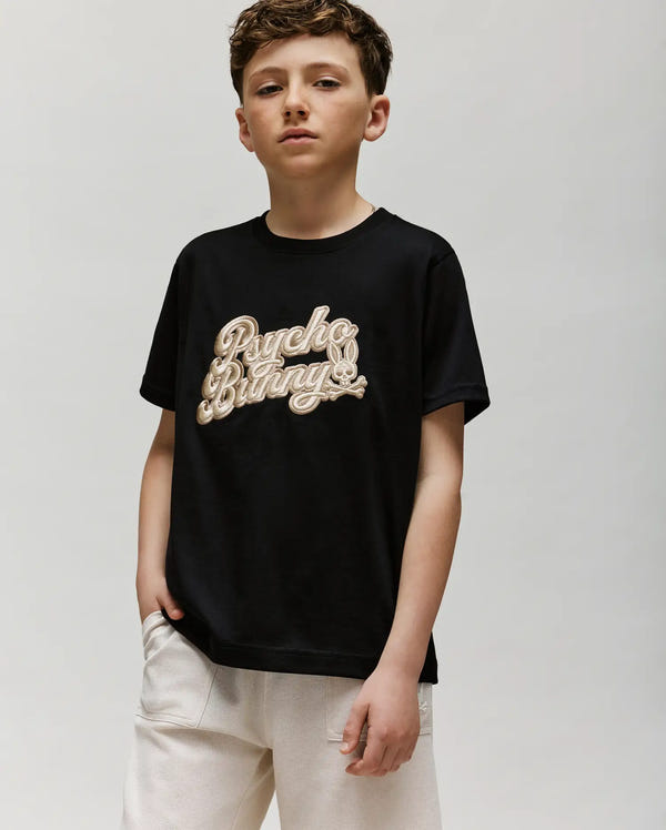 A boy with short brown hair wears a black Psycho Bunny KIDS ELIAS GRAPHIC TEE (KT0200013) made from soft Pima cotton and light shorts, standing with one hand in his pocket against a plain, light background.