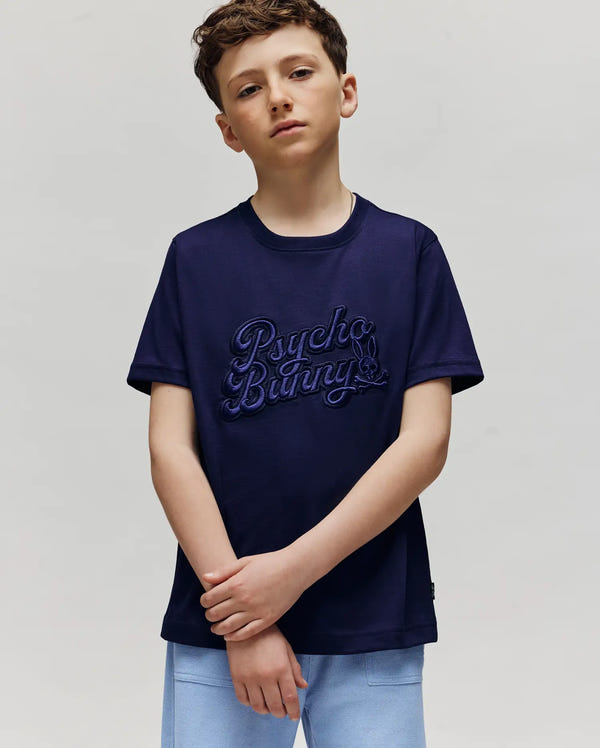 A boy with short brown hair wears the navy KIDS ELIAS GRAPHIC TEE (KT0200013) by Psycho Bunny, featuring a bold bunny graphic and light blue shorts, standing against a plain light gray background with arms crossed casually.