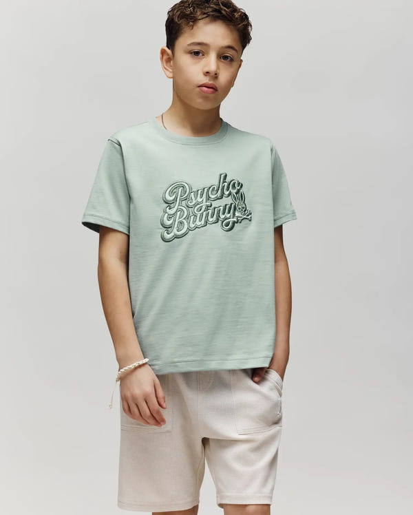 A boy with short curly hair wears the Psycho Bunny KIDS ELIAS GRAPHIC TEE (KT0200013) in light green, featuring a Bunny graphic and logo, paired with light-colored shorts. He stands with one hand in his pocket against a plain light gray background.