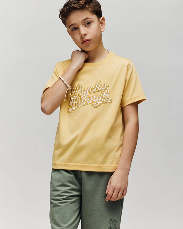 A boy wears the KIDS ELIAS GRAPHIC TEE by Psycho Bunny, featuring a bold bunny graphic in yellow, paired with green shorts. He stands against a light gray background, looking at the camera with a neutral expression.
