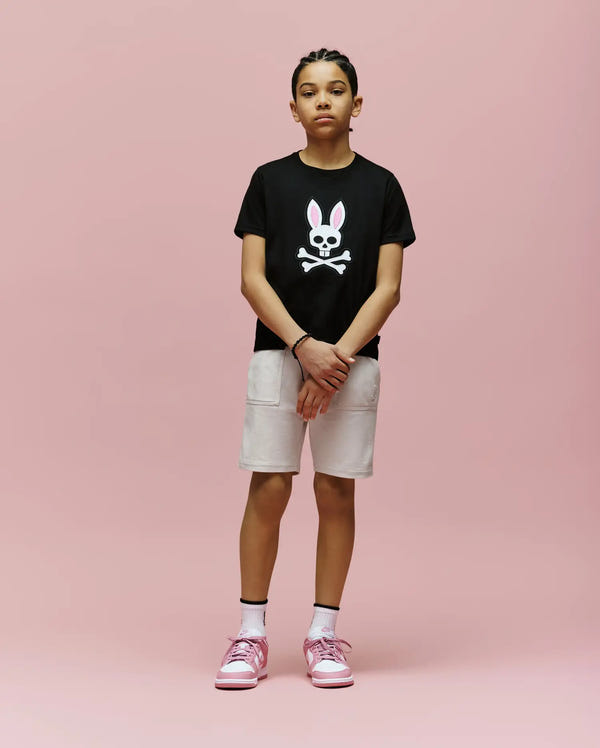 A young person stands against a pink background, wearing the Psycho Bunny KIDS ARCHIVE BUNNY GRAPHIC TEE (KT0200019) in black, paired with beige shorts, white socks, and pink sneakers. Their hands are clasped in front.