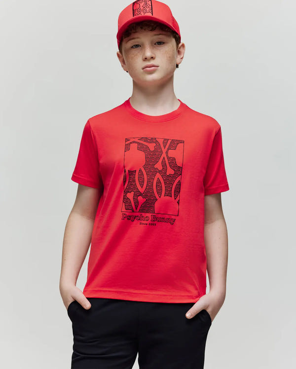 A young boy stands with hands in pockets, wearing the Psycho Bunny KIDS GLENN GRAPHIC TEE (KT0200020) in red and a matching cap. The soft Pima cotton shirt features a black abstract graphic and Psycho Bunny branding on the front.