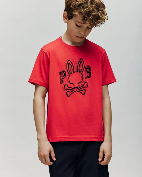 A boy with curly hair wears the Psycho Bunny KIDS BALES GRAPHIC TEE (KT0200021), a red Pima cotton shirt featuring a bold bunny skull and crossbones "P" and "B" Big Bunny print. He stands against a plain light background—a cool wardrobe addition.