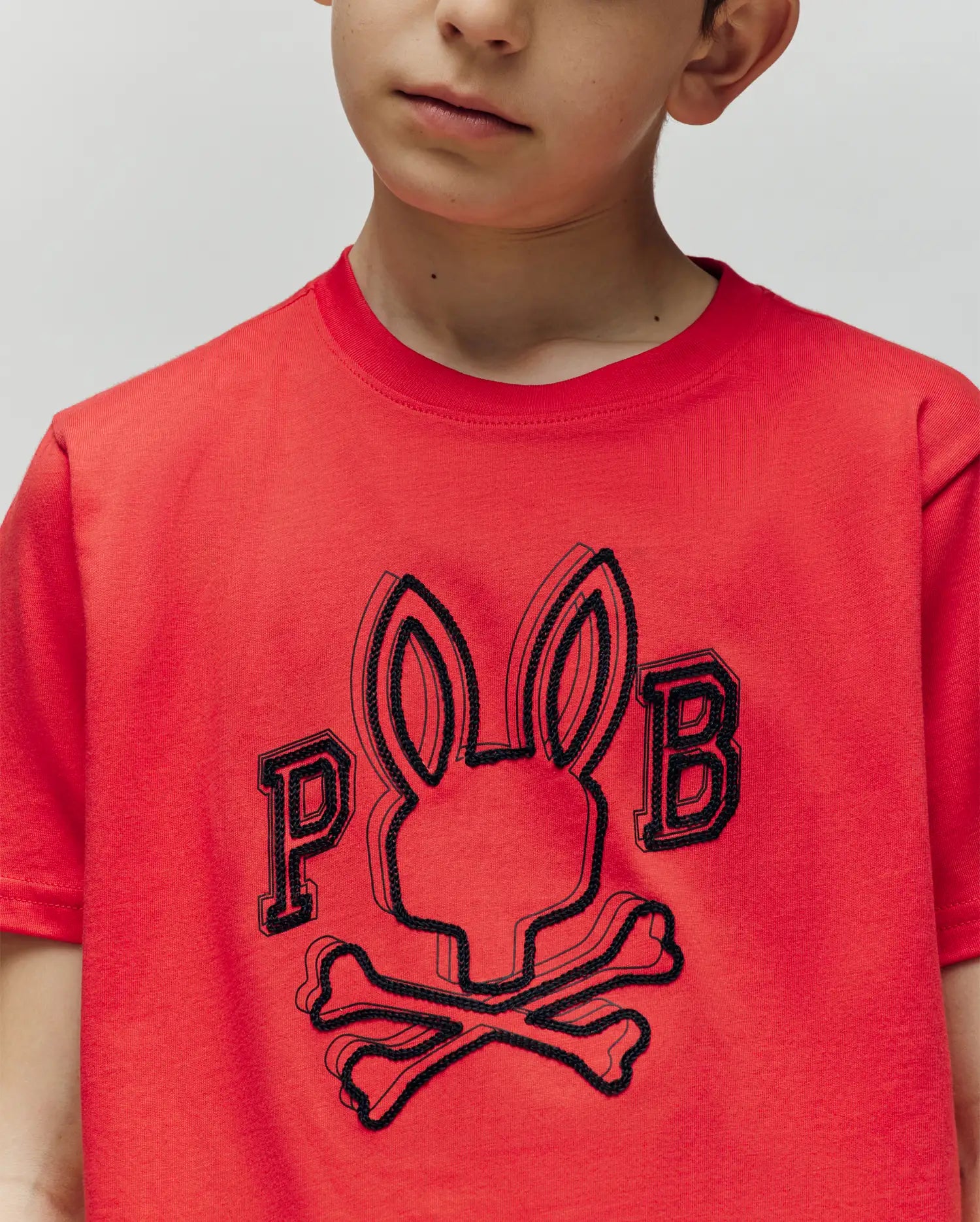 A young boy wears the Psycho Bunny KIDS BALES GRAPHIC TEE (KT0200021), a vibrant red Pima cotton shirt featuring a bold Big Bunny with crossbones and "P" and "B" graphics—perfect to brighten any wardrobe.