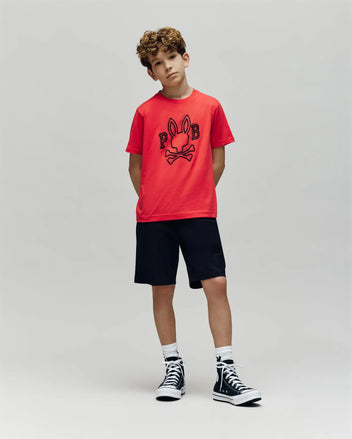 A boy with curly hair stands against a plain background wearing the Psycho Bunny KIDS BALES GRAPHIC TEE (KT0200021) in bright red, paired with black shorts, white socks, and black high-top sneakers—a standout piece for any wardrobe.