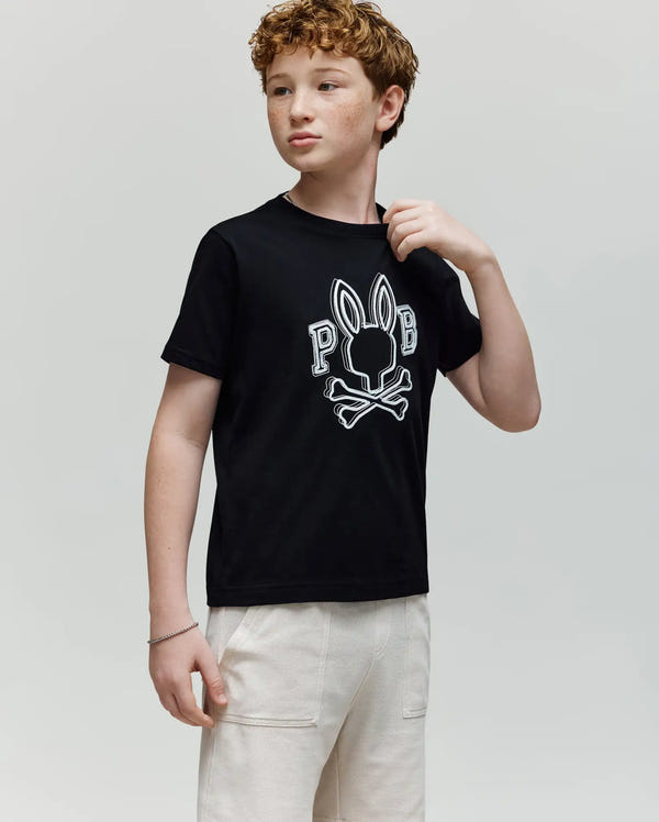 A young boy with curly red hair wears a Psycho Bunny KIDS BALES GRAPHIC TEE (KT0200021) in black featuring a Big Bunny print, paired with light-colored pants, standing against a plain background and looking slightly to the side.
