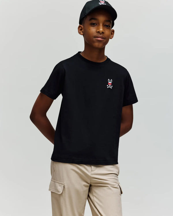 A boy stands with one hand in his pocket against a plain background, wearing the Psycho Bunny KIDS MISCHIEF BUNNY TEE (KT0200023) in black, made from soft Pima cotton with an embroidered skull and bunny logo, and beige cargo pants.