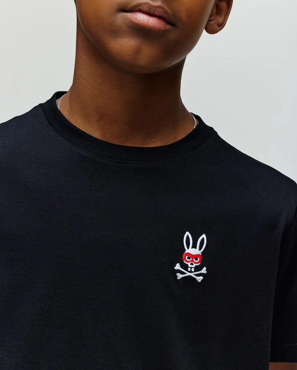 A child wearing the Psycho Bunny KIDS MISCHIEF BUNNY TEE (KT0200023)—a black Pima cotton t-shirt with a small embroidered white rabbit head, red eyes, and skull-and-crossbones—cropped to show just the lower face and upper torso.
