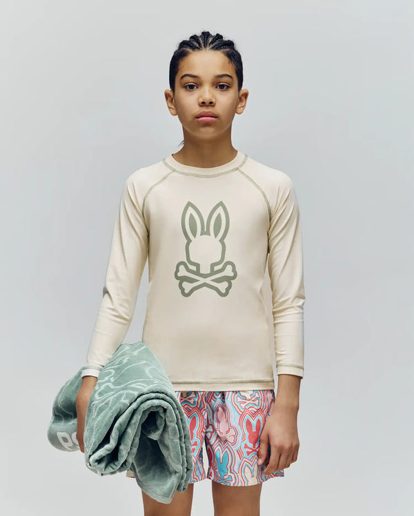A young person with braids wears the Psycho Bunny KIDS LONG SLEEVE RASHGUARD - KT0200027 in beige, featuring a bunny and crossbones graphic, paired with colorful swim trunks, holding a green towel against a plain light background.