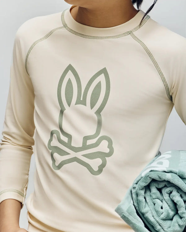 A child in a Psycho Bunny KIDS LONG SLEEVE RASHGUARD (KT0200027), featuring a large bunny and crossbones graphic and visible stitched seams, holds a rolled-up green towel. The beige shirt offers UPF 50+ UV protection with quick-drying stretch nylon.