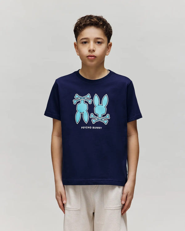A boy wears the Psycho Bunny KIDS DAWSON GRAPHIC TEE (KT0200030), a navy blue T-shirt featuring blue and white bunny and bone graphics with "Psycho Bunny" text, standing against a plain light gray background.