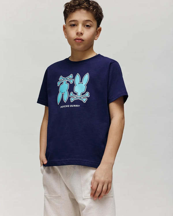 A boy with short curly hair wears the Psycho Bunny KIDS DAWSON GRAPHIC TEE (KT0200030), a navy blue shirt featuring two blue bunny graphics and "Psycho Bunny" text, paired with light pants against a plain light gray background.