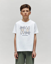 A boy with short brown hair wears a Psycho Bunny KIDS DAWSON GRAPHIC TEE (KT0200030) featuring an outline of two bunnies with crossed bones, paired with olive green shorts, standing against a plain light gray background.