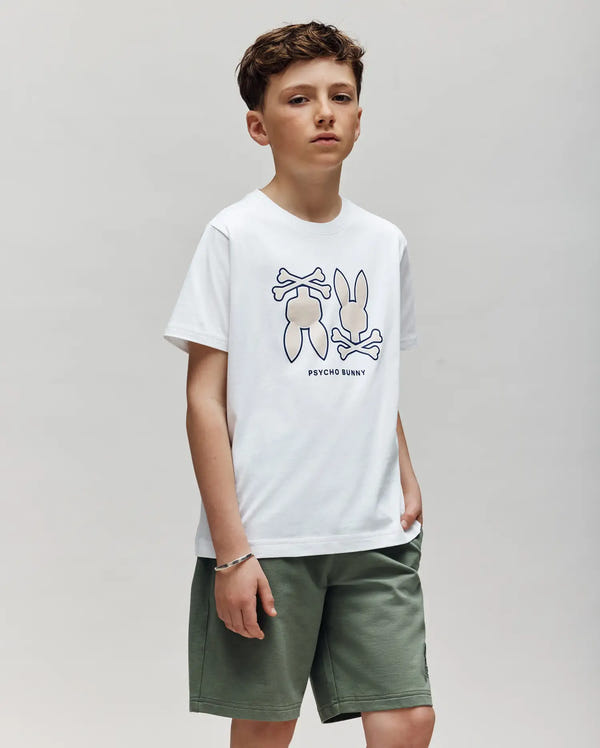 A young boy with short brown hair wears the Psycho Bunny KIDS DAWSON GRAPHIC TEE (KT0200030), a white t-shirt with cartoon bunny and crossbones graphics, paired with green shorts, standing against a plain light background.