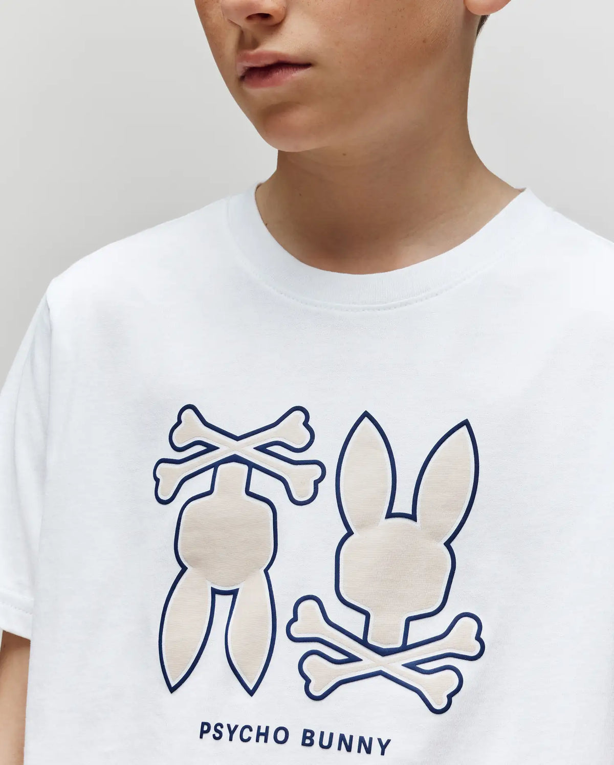 A child wears the Psycho Bunny KIDS DAWSON GRAPHIC TEE (KT0200030), a white T-shirt featuring two stylized bunny and crossbones logos with "PSYCHO BUNNY" printed beneath the graphics.