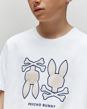 A child wears the Psycho Bunny KIDS DAWSON GRAPHIC TEE (KT0200030), a white T-shirt featuring two stylized bunny and crossbones logos with "PSYCHO BUNNY" printed beneath the graphics.