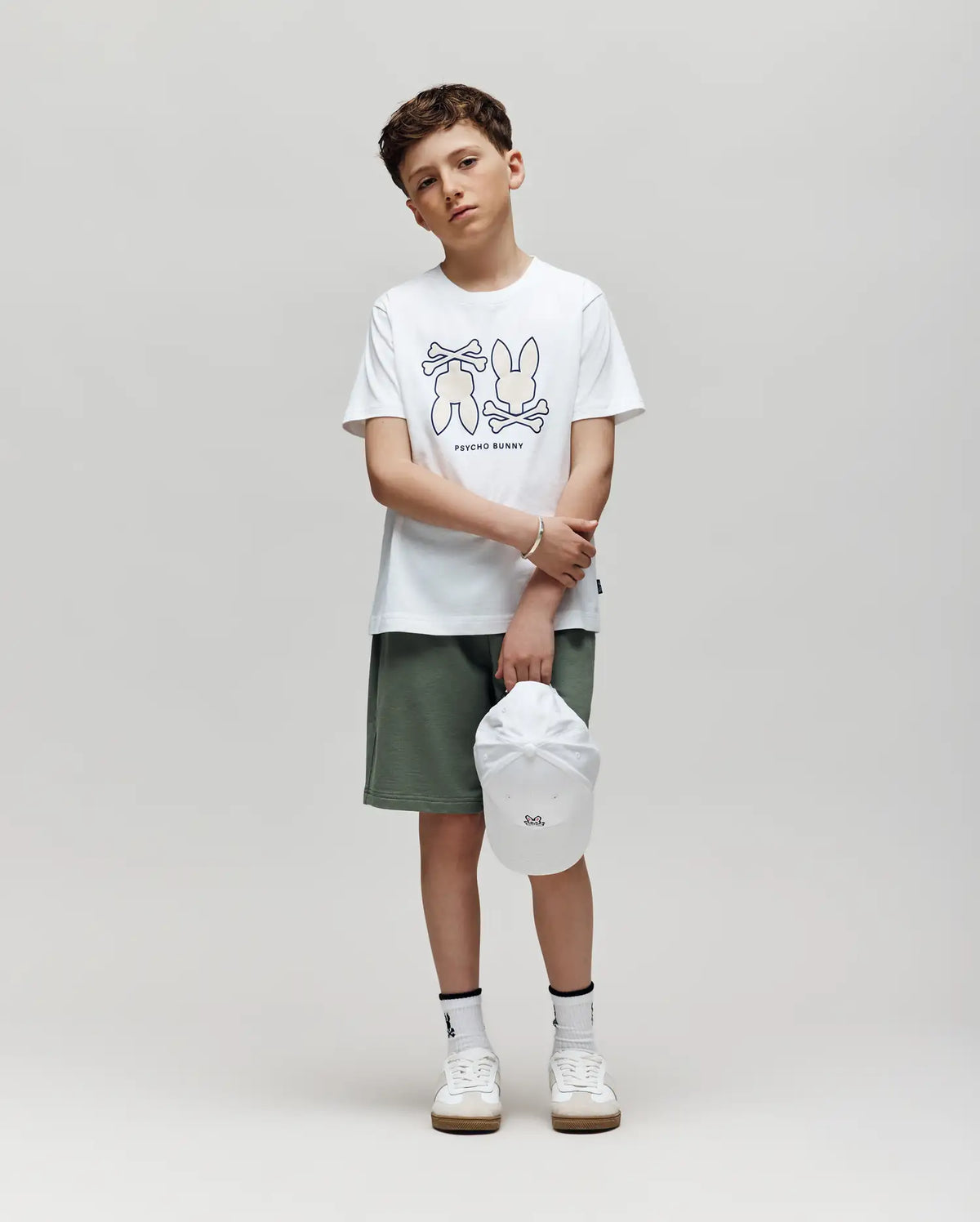 A boy stands against a plain background, wearing the Psycho Bunny KIDS DAWSON GRAPHIC TEE (KT0200030) with cartoon bunnies, green shorts, white socks, sneakers, and holding a white cap. He looks slightly serious.