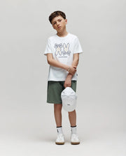 A boy stands against a plain background, wearing the Psycho Bunny KIDS DAWSON GRAPHIC TEE (KT0200030) with cartoon bunnies, green shorts, white socks, sneakers, and holding a white cap. He looks slightly serious.
