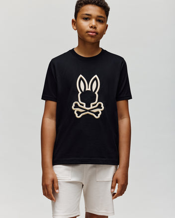 A boy in white shorts wears the Psycho Bunny KIDS NICHOLAS EMBROIDERED TEE (KT0200037), featuring the brand's signature bunny and crossbones logo, and looks at the camera with a neutral expression against a plain background.